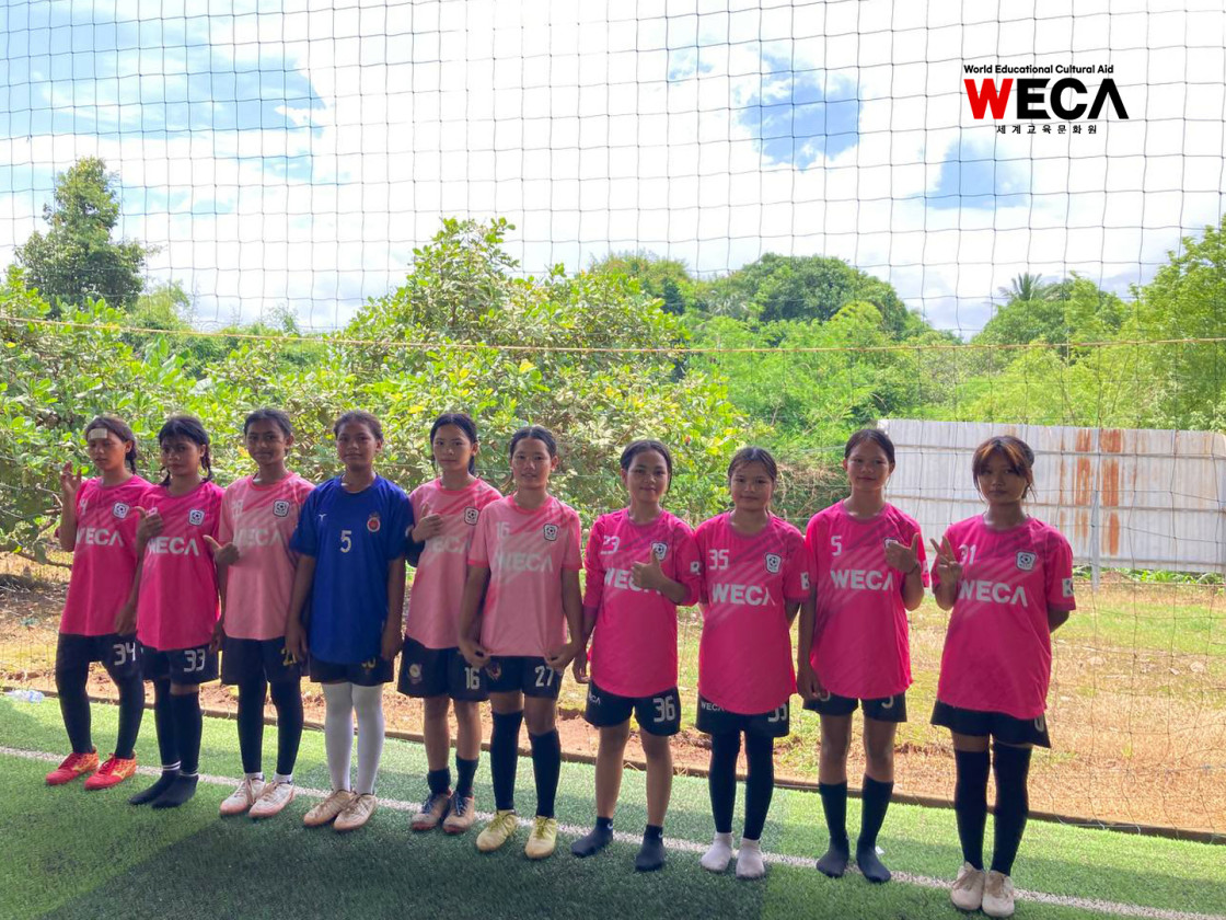 WECA의 축구 유니폼을 입고 카메라를 보고 웃으며 서있는 여자 유소년 WE-FC 축구단 아이들